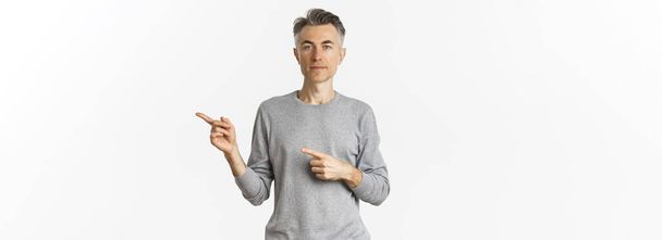 Arckép vonzó középkorú férfi szürke pulóver, mutató ujjak jobb és komoly, mutatja a reklám, áll a fehér háttér. - Fotó, kép