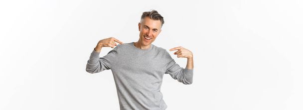 Image d'un homme beau et confiant d'âge moyen avec une coiffure grise courte, se montrant du doigt et souriant, se montrant sur fond blanc. - Photo, image