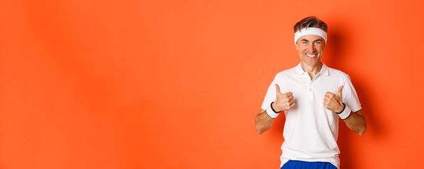Concept of sport, fitness and lifestyle. Portrait of confident, attractive middle-aged male athlete in workout uniform, showing thumbs-up in approval and smiling, orange background. - Fotoğraf, Görsel
