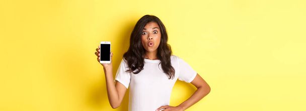 Kuva hämmästynyt afrikkalais-amerikkalainen tyttö, etsivät kiehtonut ja näyttää älypuhelin näyttö, seisoo yli keltainen tausta. - Valokuva, kuva