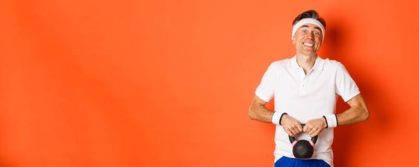 Koncepciója edzés, tornaterem és életmód. Portré vicces középkorú sportoló próbál emelni kettlebell, felett áll a narancs háttér. - Fotó, kép