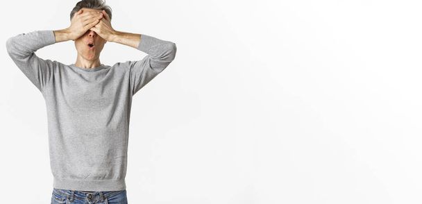 Image of excited middle-aged guy in grey sweater, cover eyes with hands and looking amazed while waiting for surprise, playing hide-n-seek, standing over white background. - Photo, Image