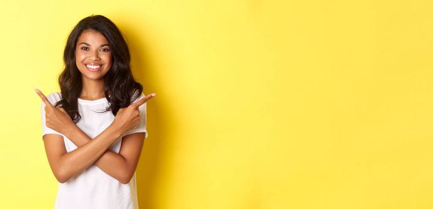 Retrato de menina afro-americana feliz e confiante mostrando duas escolhas, apontando os dedos para o lado direito e esquerdo espaço de cópia, sorrindo satisfeito, em pé sobre fundo amarelo. - Foto, Imagem