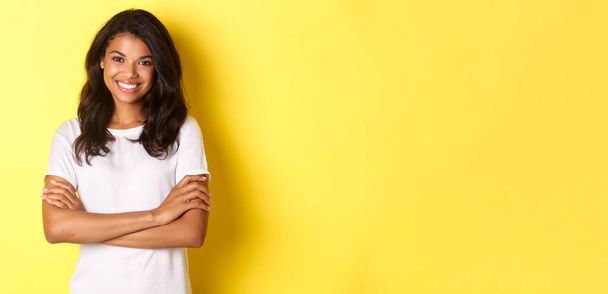 Image of happy smiling african-american girl in white t-shirt, cross arms on chest and looking confident, standing over yellow background. - Фото, изображение
