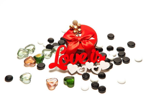 Red round box with a bow and decoration on top, the inscription love is, white and black round shiny stones, glass hearts of different colors on a white background. - Photo, Image
