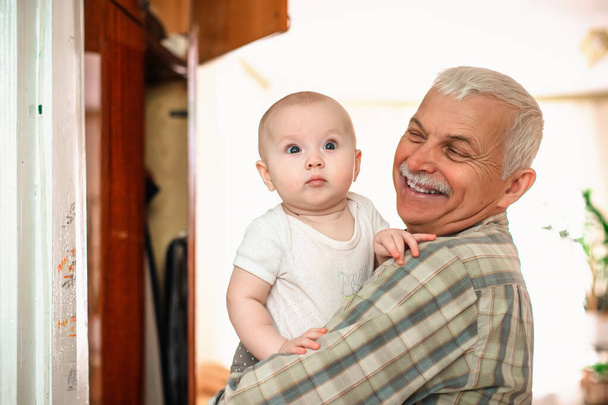 Szczęśliwy uśmiechnięty dziadek bawi się ze swoim wnuczkiem w pokoju przy oknie. Starzec i dziecko w wieku do jednego roku - Zdjęcie, obraz