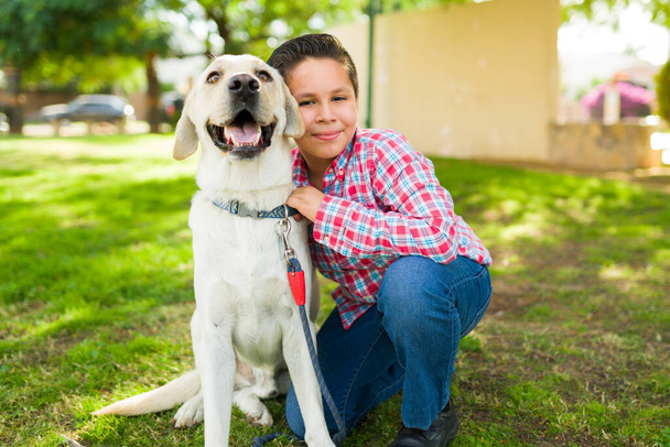 Parkta oynarken labrador köpeğine gülümseyen ve sarılan sevimli Latin bir çocuğun portresi. - Fotoğraf, Görsel