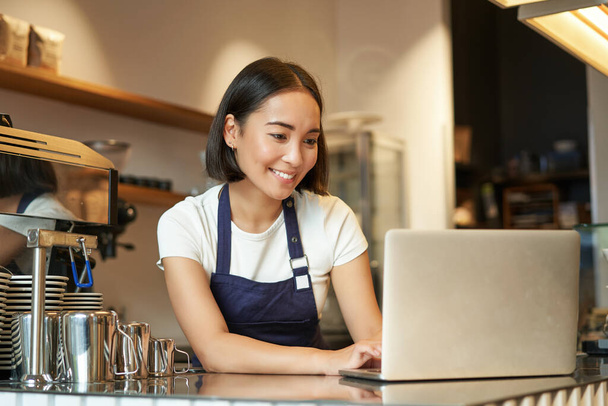 Ritratto di barista asiatica sorridente, imprenditore proprietario di caffè, lavorando su laptop, elaborando ordini su computer, in piedi dietro il bancone. - Foto, immagini