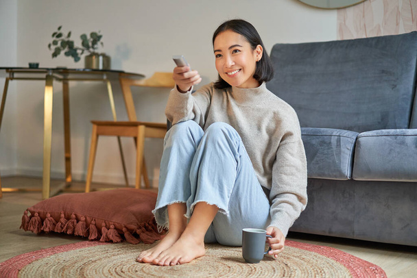 Portré gyönyörű ázsiai lány ül az otthonában, és TV-t néz, kezében távoli, mosolygós és nevető, érzés kényelem és melegség a lakásában. - Fotó, kép