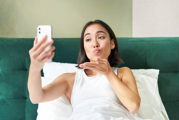Linda menina asiática deitada na cama, fazendo selfie manhã, tirando foto no smartphone no quarto, sorrindo feliz. - Foto, Imagem