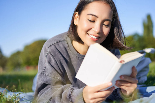 Egy gyönyörű, mosolygó ázsiai lány portréja, aki a parkban olvas, a fűben fekszik kedvenc könyvével. Szabadidő és emberek koncepciója. - Fotó, kép