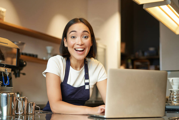 Ενθουσιασμένη Ασιάτισσα με στολή καφετέριας, μπαρίστα με φορητό υπολογιστή, να δείχνει χαρούμενη και έκπληκτη με την κάμερα. - Φωτογραφία, εικόνα