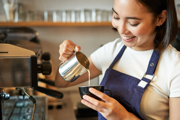 Smiling asian barista girl, making coffee, pouring steamed milk into cappuccino, doing latte art in cup, working in cafe. - Photo, Image