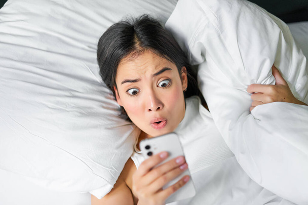 Portret van aziatisch meisje wakker in de ochtend, kijkt naar mobiele telefoon met geschokt gezicht, verslapen, knuffelen kussen en grimmig. - Foto, afbeelding