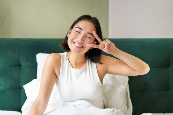 Pozitif Asyalı kadın yatakta uzanıyor, barış işareti gösteriyor, mutlu bir sabah geçiriyor, neşeli bir şekilde uyanıyor, yatak odasında kalıyor.. - Fotoğraf, Görsel