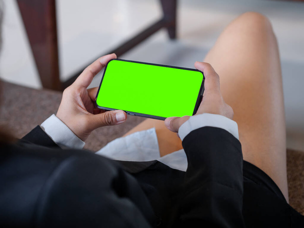 Pantalla verde en el estilo horizontal del teléfono móvil en las manos de las mujeres de negocios que usan traje formal y pantalones cortos, se sientan relajados con piernas cruzadas en el sofá en la acogedora sala de estar. Smartphone de pantalla simulada verde. - Foto, Imagen