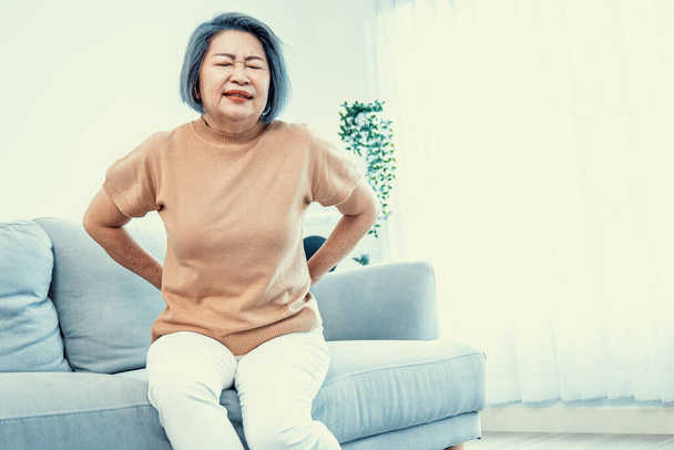 An agonizing senior woman experiencing back pain while sitting on her sofa at home and holding her back. - Photo, image