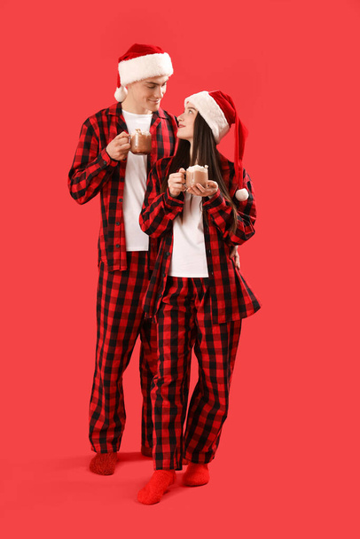Junges Paar in Weihnachtsmützen mit Tassen Kakao auf rotem Hintergrund - Foto, Bild