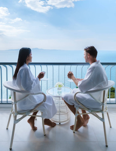 Pari aasialaista naista ja valkoihoista miestä juo kahvia hotellihuoneessa katselemassa merelle Thaimaassa. - Valokuva, kuva