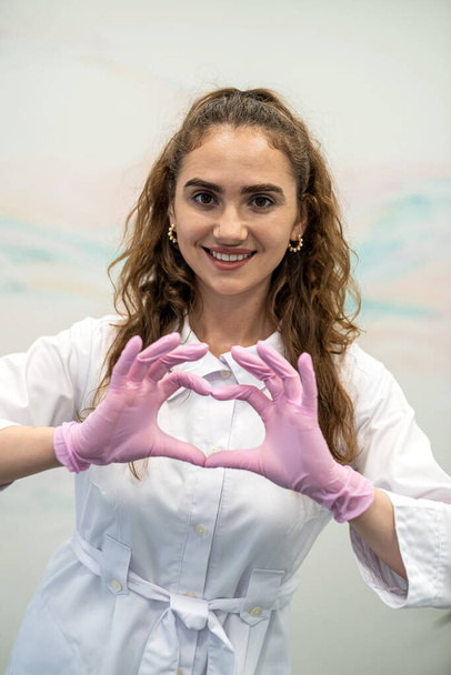Portrait d'une jeune femme médecin heureuse en uniforme médical montrant un geste ou un signe d'amour cardiaque. Infirmière souriante partageant les soins et le soutien des patients. - Photo, image