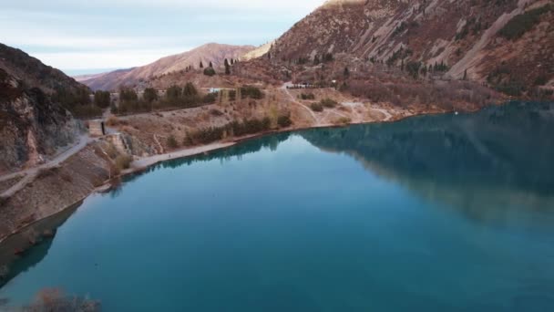 Bovenaanzicht op het Issyk bergmeer en de dam. Een riviertje stroomt het meer in. Er staan geel-groene bomen op de heuvels. Water reflecteert de lucht. Mensen lopen langs de kust en paden. Kazachstan - Video