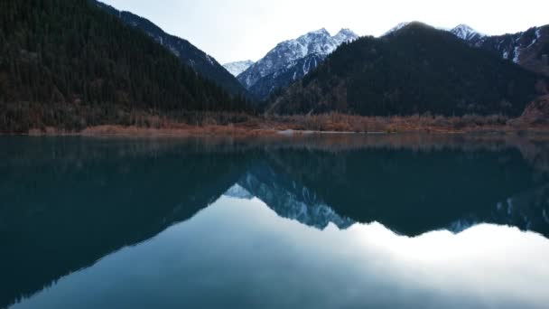 Issyk lago de montanha com espelho de água ao pôr do sol. A cor da água muda diante dos nossos olhos. Há árvores em águas claras. Montanhas nevadas e colinas verdes são visíveis. Nuvens são refletidas - Filmagem, Vídeo