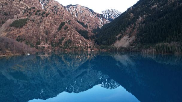 Espejo azul oscuro de agua en un lago de montaña. La superficie lisa es como un espejo, los árboles, las colinas amarillo-verdes, las montañas y el cielo se reflejan. Los troncos de los árboles están en el agua. Issyk. - Foto, imagen