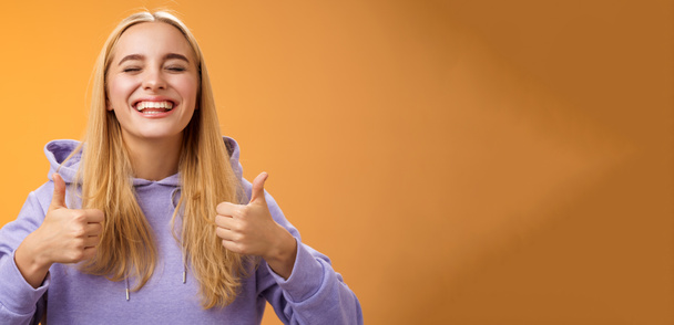 Close-up zorgeloos blij positief geluk jonge blonde vrouw in hoodie sluit ogen vrolijk vieren goed resultaat show duimen-up gebaar eens goedkeuring geweldig idee, oranje achtergrond. - Foto, afbeelding