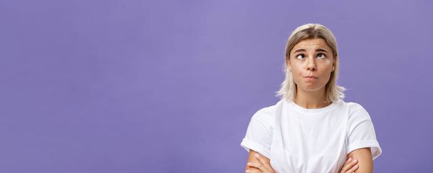 Mind going crazy. Portrait of funny tired attractive young woman with fair hair and tanned skin squinting rolling eyes aside and looking up fooling around over purple background from boredom. Emotions - Photo, Image