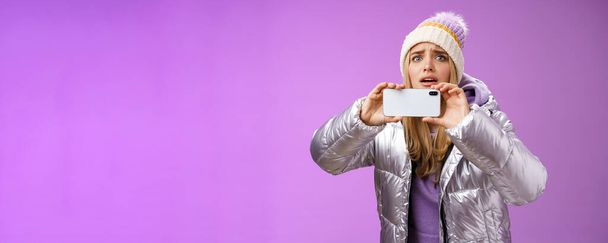 Intense worried blond girl holding mobile phone horizontal recording video capture moment share friends internet blog using smartphone camera shoot photograph standing serious purple background. - Photo, Image