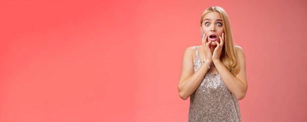 Shocked concerned stunned young european blond woman in silver dres drop jaw widen eyes stunned speechless gasping open mouth touch face amazed, look troubled perplexed, red background. - Photo, Image