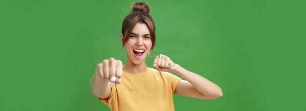 Leuke vrouwelijke rebel in geel t-shirt met gap tanden trekken vuist in de richting van de camera alsof het tonen van vechten vaardigheden schreeuwen gedurfd en opgewonden staan over groene achtergrond glimlachen gedragen als bokser. Lichaam - Foto, afbeelding