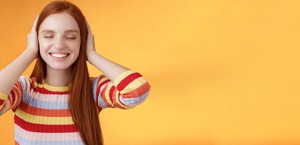 Dreamy charismatic relaxed tender redhead caucasian girl 20s close eyes cover ears imaging far away, smiling broadly enjoy silence peace comfort without sounds noise-free, orange background. - Photo, Image