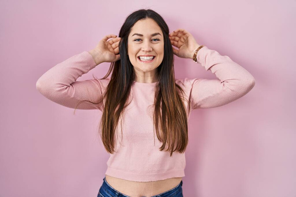 Jonge brunette vrouw staat over roze achtergrond glimlachende trekken oren met vingers, grappig gebaar. auditieprobleem  - Foto, afbeelding