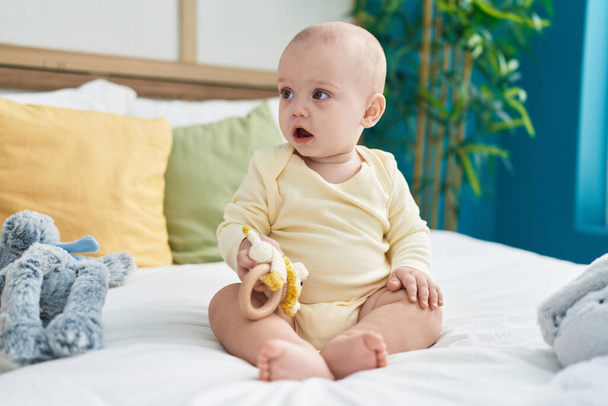 Adorable bebé caucásico sosteniendo juguete sentado en la cama en el dormitorio - Foto, Imagen