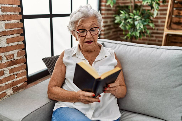 Ηλικιωμένη γκριζομάλλα γυναίκα που διαβάζει τη Βίβλο καθισμένη στον καναπέ στο σπίτι - Φωτογραφία, εικόνα