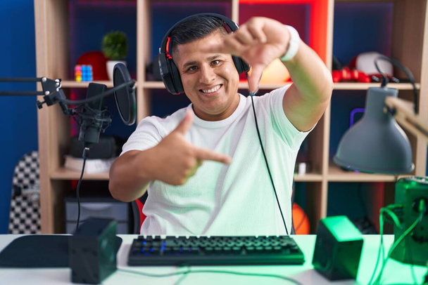 Jonge Spaanse man die videospelletjes speelt met een glimlachende lijst met handen en vingers met een vrolijk gezicht. creativiteit en fotografie.  - Foto, afbeelding