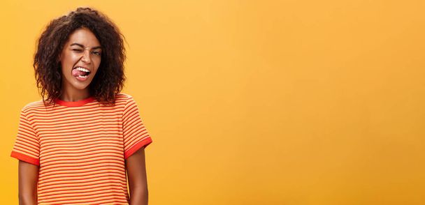 Πορτρέτο τολμηρή και συναισθηματική αυτοπεποίθηση flirty γυναίκα με afro hairstyle κλείσιμο του ματιού χαρούμενη εμφάνιση γλώσσα θέτοντας ανέμελη και ενθουσιώδης κατά πορτοκαλί φόντο φλερτ με καυτό άντρα. Αντιγραφή χώρου - Φωτογραφία, εικόνα