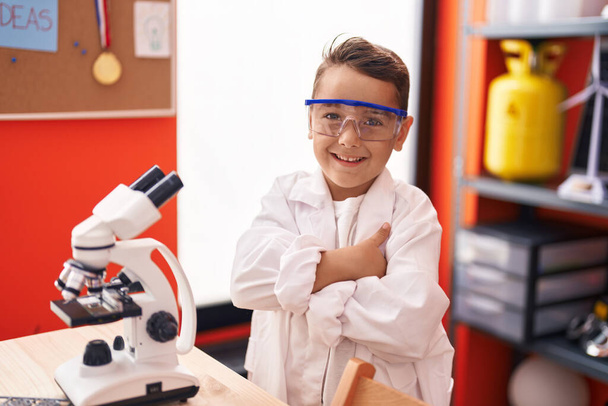 Suloinen latino lapsi opiskelija käyttäen mikroskooppi seisoo kädet ristissä ele luokkahuoneessa - Valokuva, kuva