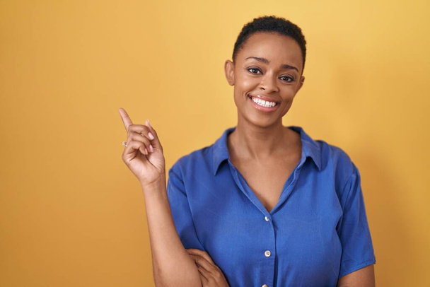 Mujer afroamericana de pie sobre fondo amarillo con una gran sonrisa en la cara, señalando con el dedo de la mano al lado mirando a la cámara.  - Foto, imagen