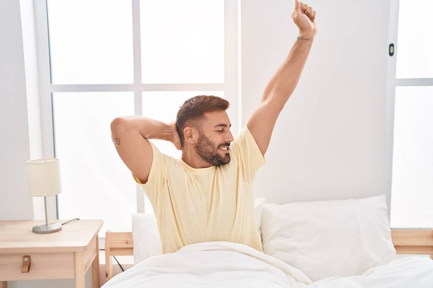 Young hispanic man waking up stretching arms at bedroom - Photo, image