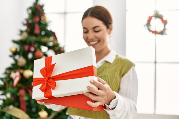 Giovane bella donna ispanica disimballaggio regalo in piedi da albero di Natale a casa - Foto, immagini