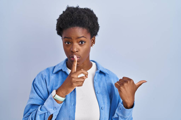 Mujer afroamericana de pie sobre fondo azul pidiendo estar callada con el dedo en los labios apuntando con la mano hacia un lado. silencio y concepto secreto.  - Foto, imagen