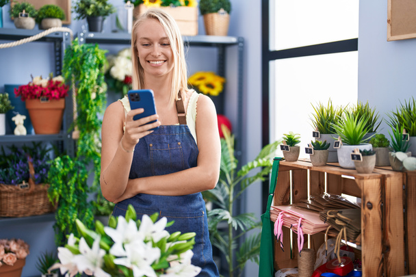 Молодая блондинка-флористка уверенно улыбается, используя смартфон в цветочном магазине - Фото, изображение