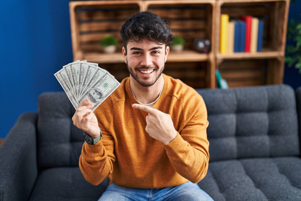 Spaanse man met baard die bankbiljetten van 100 dollar vasthoudt, glimlachend vrolijk wijzend met hand en vinger.  - Foto, afbeelding
