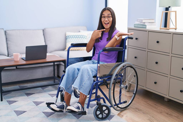 Νεαρή Ισπανίδα που κάθεται σε αναπηρική καρέκλα στο σπίτι έκπληκτη και χαμογελώντας στην κάμερα ενώ παρουσιάζει με το χέρι και δείχνοντας με το δάχτυλο.  - Φωτογραφία, εικόνα