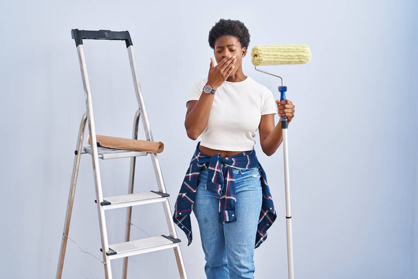 Mujer afroamericana sosteniendo rodillo pintor aburrido bostezo cansado cubriendo la boca con la mano. inquietud y somnolencia.  - Foto, imagen