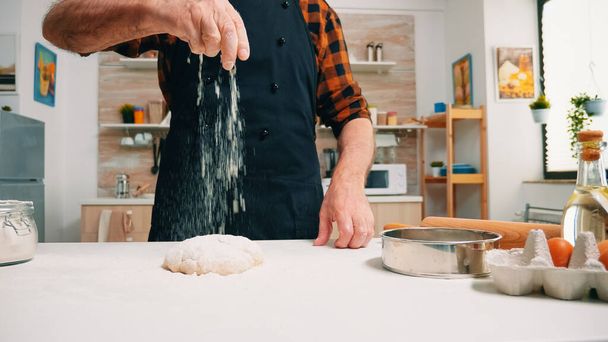 El primer plano del hombre tamizar la harina en la masa de pan en la mesa de madera en la cocina. Mayores panadero senior con bonete y uniforme de aspersión, tamizado, la propagación de los ingredientes de rew en la masa, hornear pizza casera - Foto, imagen