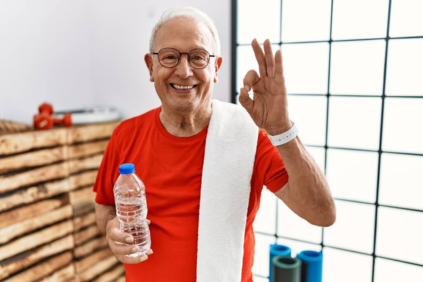 Старший мужчина в спортивной одежде и полотенце в тренажерном зале улыбаясь позитивно делает хорошо знак с рукой и пальцами. успешное выражение.  - Фото, изображение