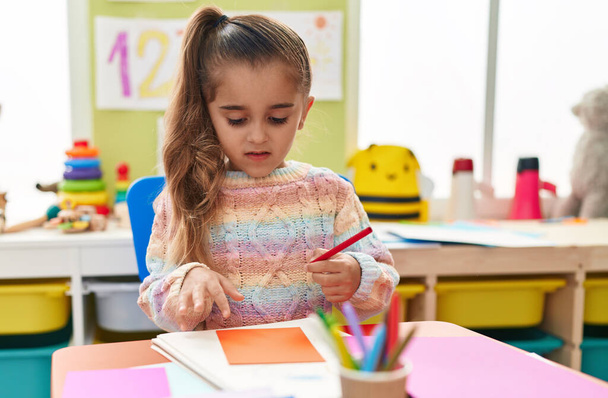 Entzückende hispanische Studentin sitzt im Kindergarten auf einem Tisch und zeichnet auf Papier - Foto, Bild